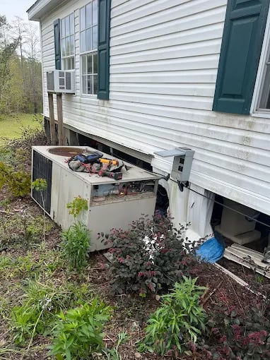 Old air conditioning unit with debris sits beside weathered white house