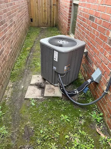 Gray air conditioning unit installed beside brick wall with overgrown plants